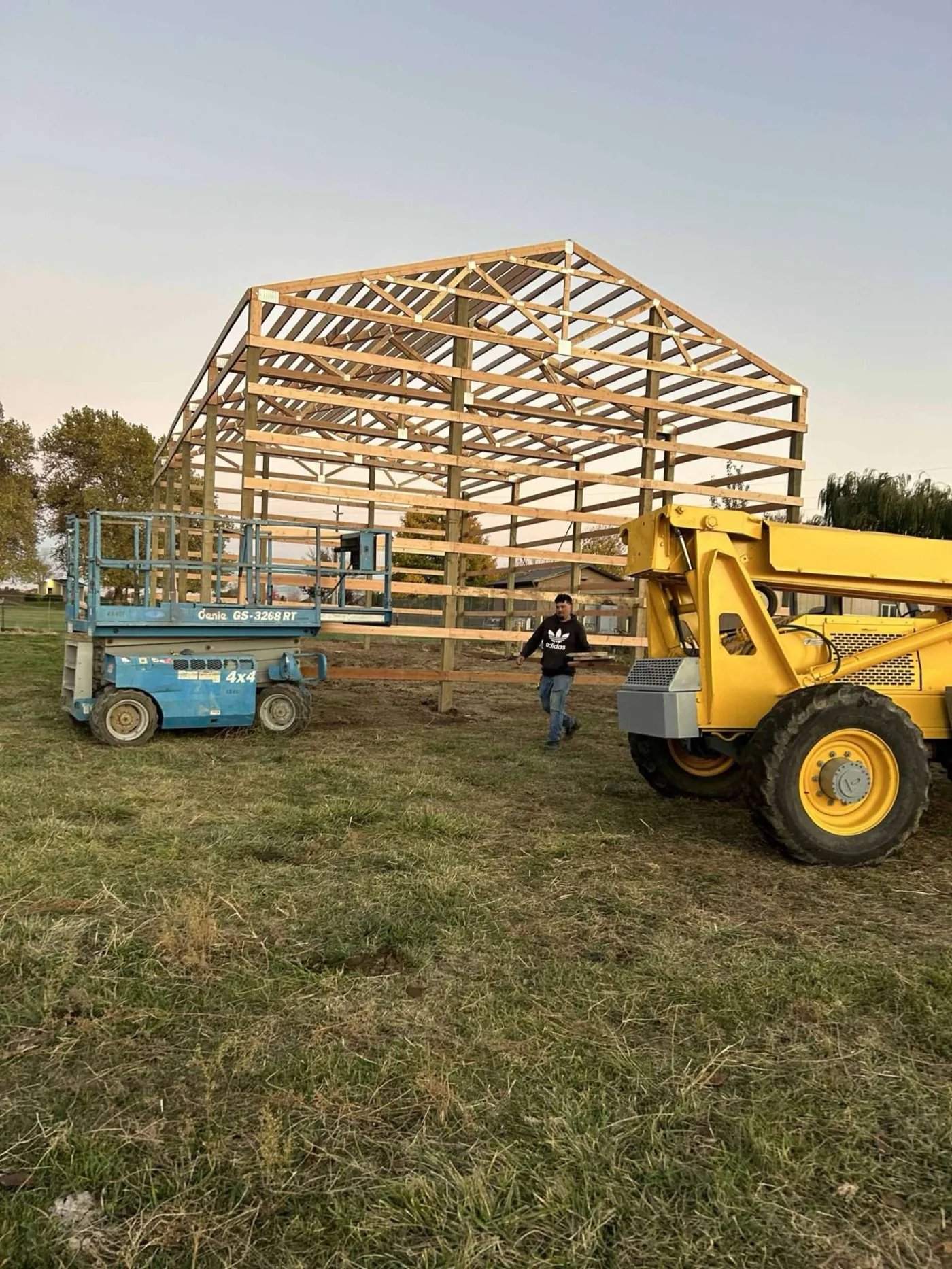 Crew working on a pole building frame at dusk with a reach forklift and scissor lift on site