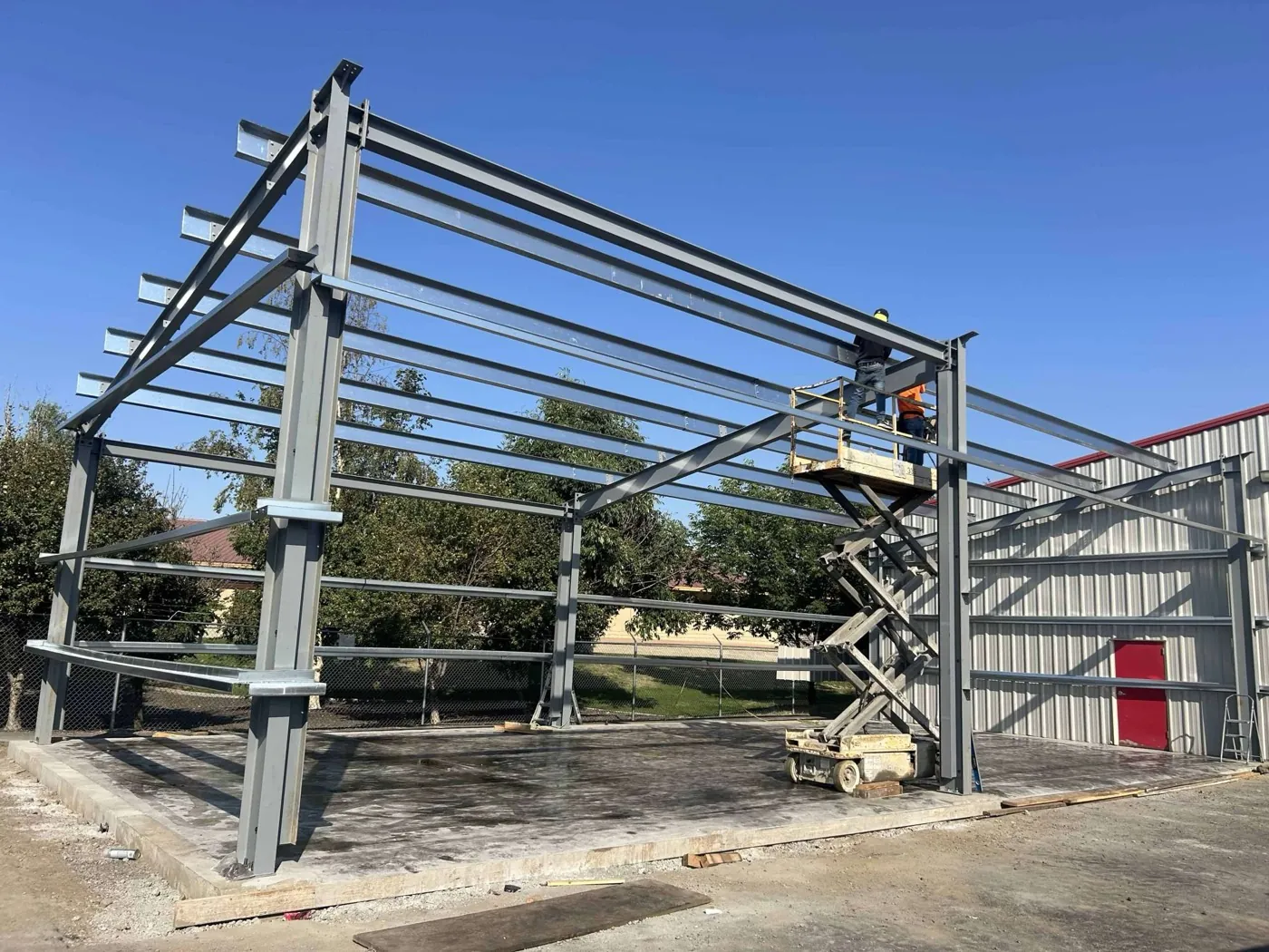Crew on a scissor lift assembling a steel building frame, an unrelated finished building visible behind it
